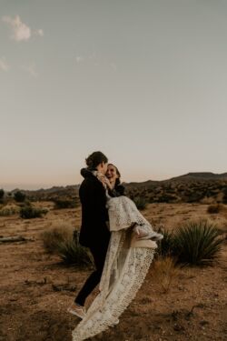 Allison wearing Lisa Dress | Randi Kreckman Photography joshua-tree-wedding-bride-swept-up-by-her-groom-wearing-backless-bohemian-lace-dress-with-pink-converse